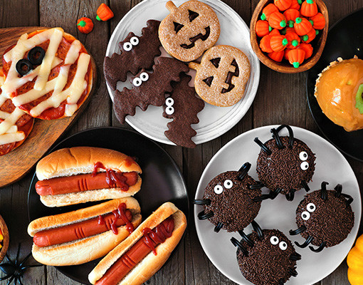 Halloween party food table scene over a rustic wood background. Overhead view. Spooky mummy pizzas, finger hot dogs, caramel apples, cupcakes, candy, cookies and donuts.